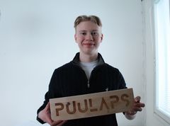 Henkilö pitelee puista kylttiä, jossa lukee "PUULAPS".