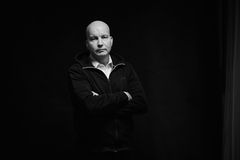 Tummataustainen mustavalkovalokuva henkilöstä, joka seisoo käsivarret ristissä vakavana.