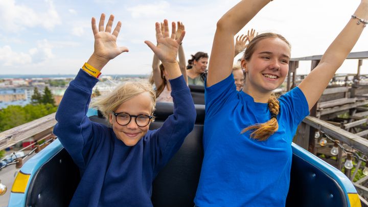 Kaksi hymyilevää nuorta puisen Vuoristoradan kyydissä, kädet ylhäällä.