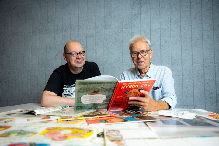 Mauri Kunnas ja Harri Römpötti lukevat Nyrok City -sarjakuvaa pöydän ääressä.
