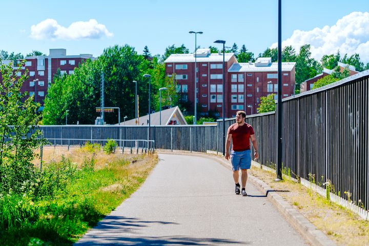 Henkilö kävelee kevyenliikenteen väylällä. Väylän oikeassa reunassa on puinen meluaita. Taustalla näkyy punainen kerrostalo.