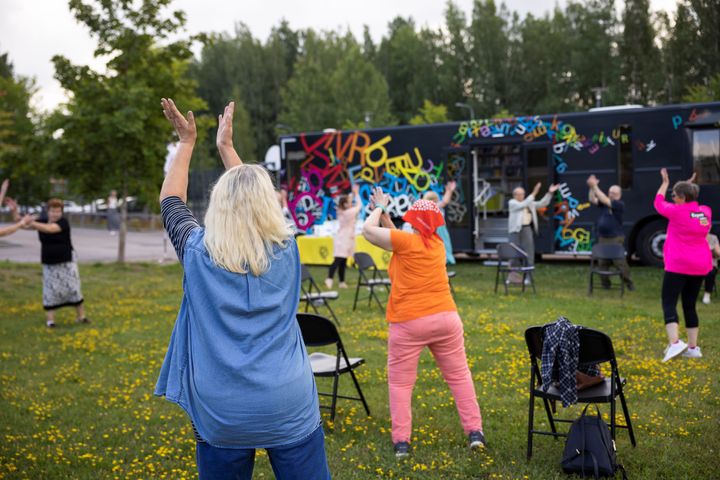 Ihmisiä jumppaamassa ulkona nurmikolla kesäpäivänä. Taustalla näkyy Senioripaku Välkky.