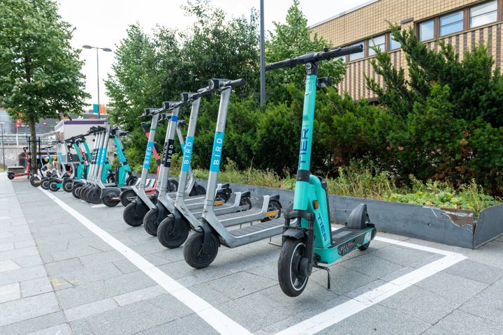 Monenvärisiä sähköpotkulautoja pysäköityinä toimistorakennuksen edessä.