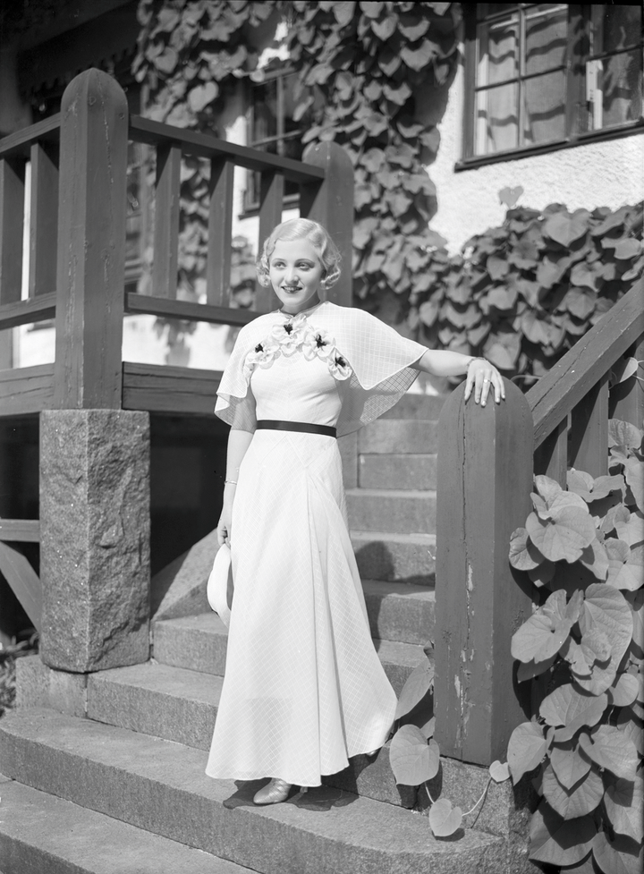 Mustavalkoisessa kuvassa nainen valkoisessa mekossa seisomassa rakennuksen portailla.