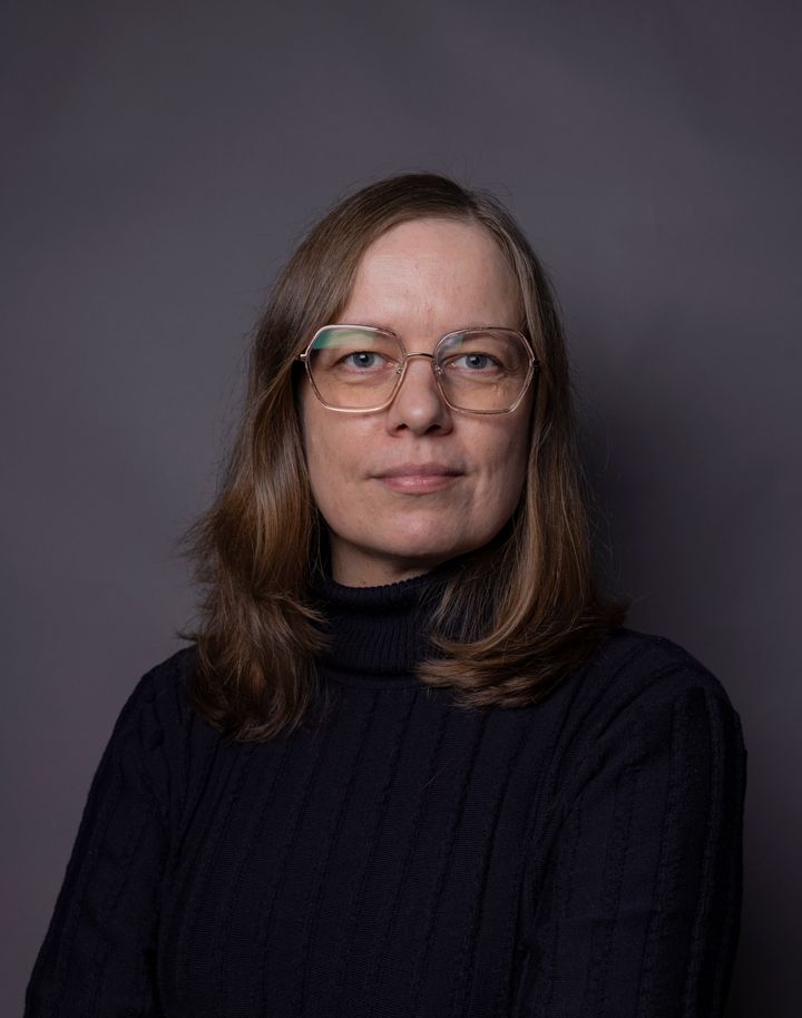 Potrettikuva silmälasipäisestä naisesta tummassa vaatteessa, tummaa taustaa vastaan.