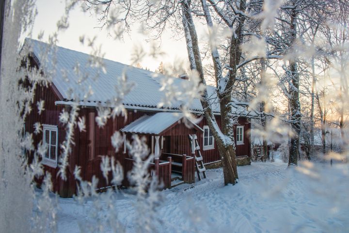 Lumiessa maisemassa punainen vanha puutalo talvisen auringonlaskun aikaan.