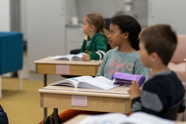 Kolme koululaista istuu pulpeteissa luokkahuoneessa oppikirjat avoinna.
