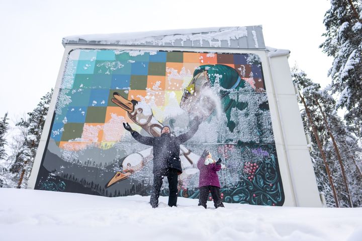 Henkilöt leikkivät lumessa värikkään muraalin edessä talvisessa maisemassa.