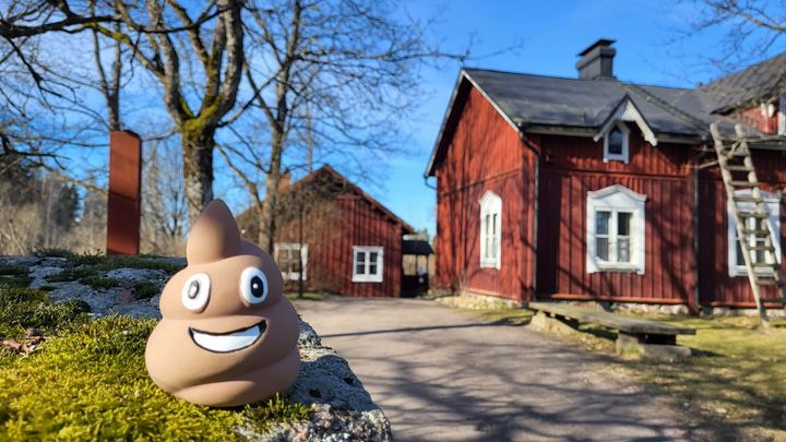 Iso kakkaemojin näköinen patsas punaisten rakennusten, Talomuseo Glimsin edessä,  portin pielessä aurinkoisena kevätpäivänä kuvattuna.