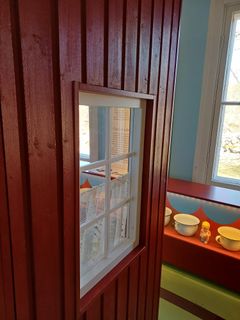 Punaisen huussin seinä, jossa on valkoinen ikkuna ja porsliinipottia taaempana hyllyllä.