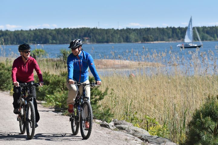 Kaksi pyöräilijää ajavat hiekkatietä pitkin, taustalla näkyy meri ja purjevene.