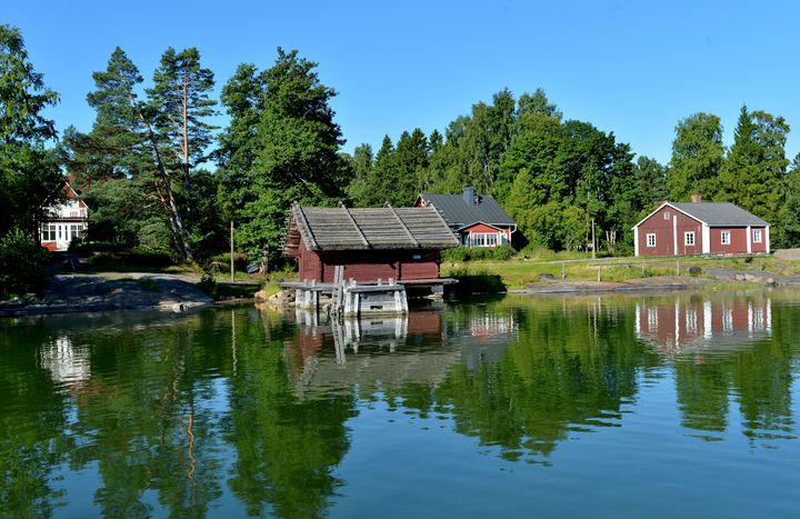 Museoalue kuvattu mereltä päin, kolme punaista puutaloa vihreiden lehtipuiden lomassa.