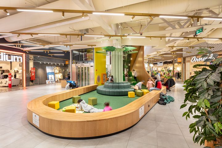 Lasten leikkipaikka kauppakeskuksen sisällä, jossa on vieressä liikkeitä ja viherkasvi.
