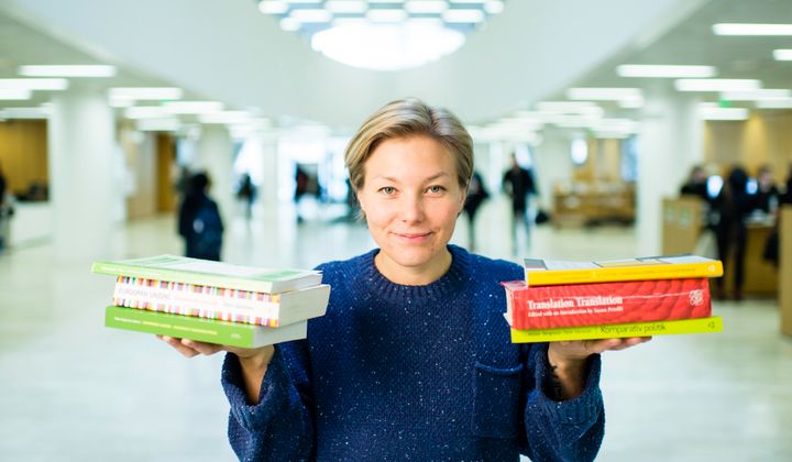 Mia Pohtola seisoo kirjastossa ja pitelee kummallakin kämmenellään pinoa kirjoja.