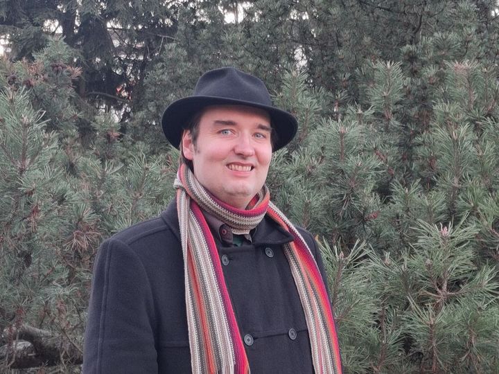 Petri Tikka seisoo metsämaisemaa vasten hymyillen, yllään musta takki, hattu ja raidallinen kaulaliina.