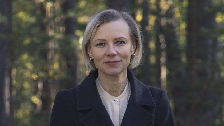 Jonna Alava seisoo metsäisen maiseman edessä hymyillen ja katsoo kameraan.