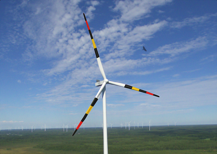 Dronekuva tuulivoimalasta Etelä-Suomessa. Voimalan lavat on kuvioitu värikkäin varoituskuvioin. Taustalla näkyy lisää tuulivoimaloita ja avara maisema.