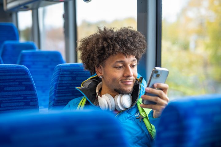 A person with curly hair and headphones looks at a smartphone while sitting on a bus.