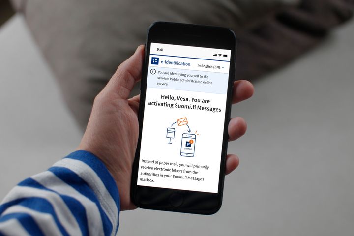 In the picture, a person is holding a phone in their hand, and the phone screen displays a notification about the launch of Suomi.fi messages.