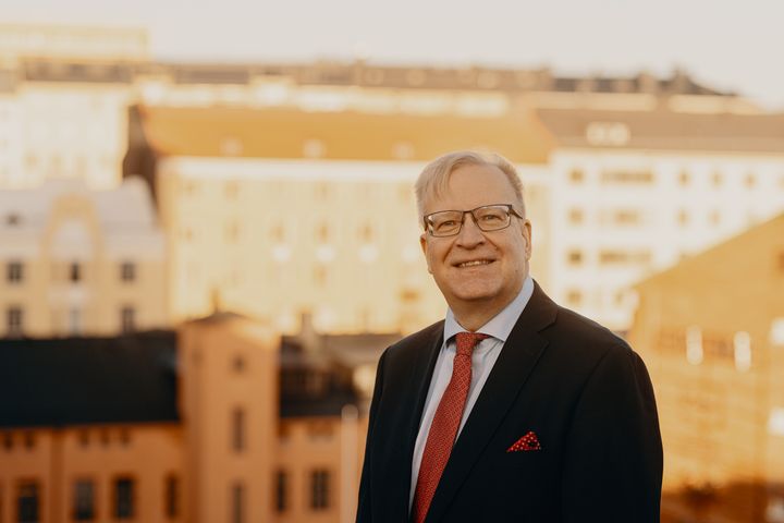 Digi- ja väestötietoviraston Kimmo Rousku seisoo kattojen yläpuolella kaupunkimaisemassa pukeutuneena pukuun ja solmioon.