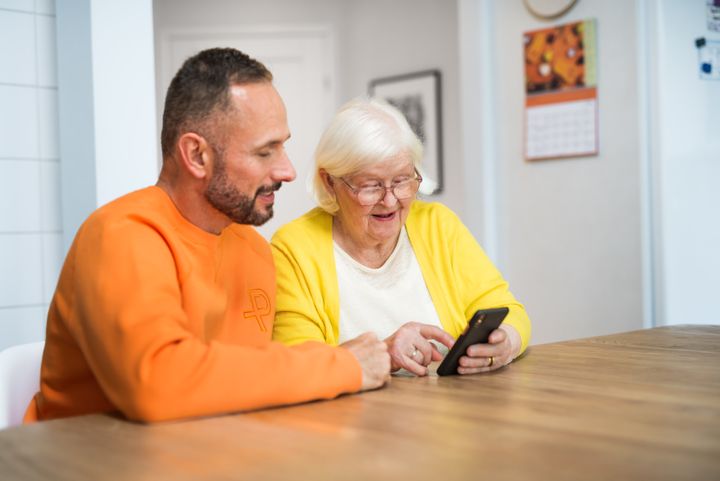 Kuvassa kaksi henkilöä istuu pöydän ääressä katsoen yhdessä älypuhelinta.