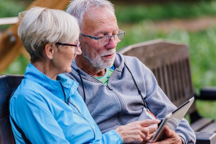 Äldre par sitter på en bänk utomhus och tittar på en surfplatta tillsammans.