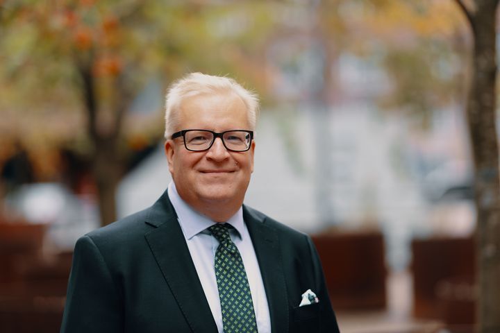 Kimmo Rousku seisoo pihalla ja hymyilee kameraa kohti. Kimmon yllä on tumma puku. Hänen taustallaan näkyy sumeana puita.