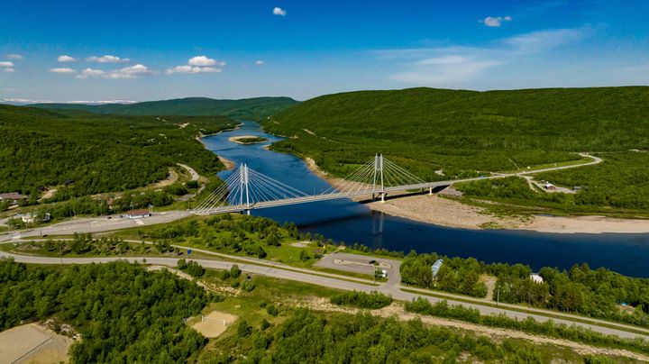 Flygbild av en flod som rinner genom ett somrigt fjällandskap. En vajerbro går över floden.