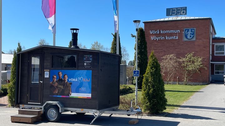 Musta siirreltävä mökki pysäköitynä parkkipaikalle, taustalla näkyy Vöyrin kunnantalo ja suurikokoinen kello näyttää aikaa 13:36.