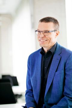 Kuvassa on hymyilevä henkilö, jolla on sininen puku ja silmälasit.