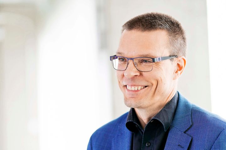 Kuvassa on hymyilevä henkilö, jolla on sininen puku ja silmälasit.