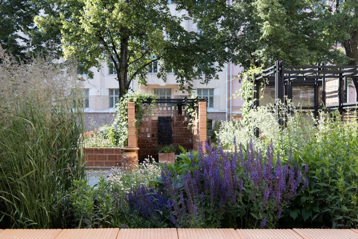 Savesta ja ekologisista materiaaleista rakennettu paviljonki ulkoilmassa, paljon vihreää kasvillisuutta sen ympärillä