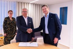 Kolme henkilöä toimistossa, kaksi miestä kättelee neuvottelupöydän ääressä, kolmas henkilö seisoo taustalla hymyillen. Pöydällä on asiakirjoja.