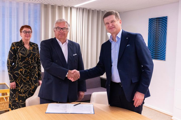 Kolme henkilöä toimistossa, kaksi miestä kättelee neuvottelupöydän ääressä, kolmas henkilö seisoo taustalla hymyillen. Pöydällä on asiakirjoja.