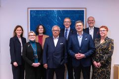 Ryhmä ihmisiä seisoo rivissä ja hymyilee kameralle. He ovat pukeutuneet siisteihin vaatteisiin, ja taustalla on sininen taideteos.