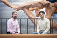 Kaksi henkilöä istuu metallikaiteen edessä, taustalla ruskea veistos.