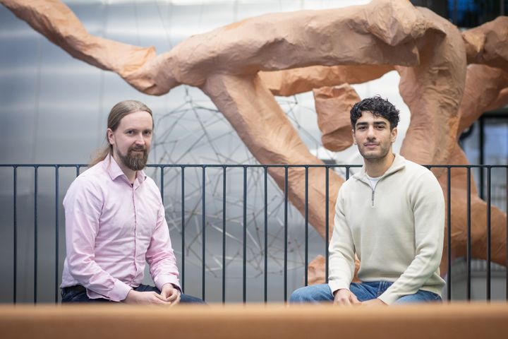 Kaksi henkilöä istuu metallikaiteen edessä, taustalla ruskea veistos.