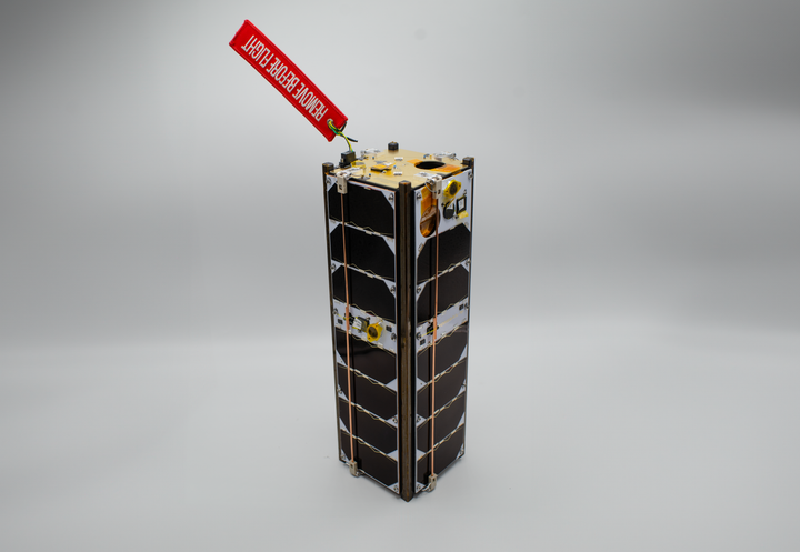 A small satellite with solar panels and a red "Remove Before Flight" tag attached.