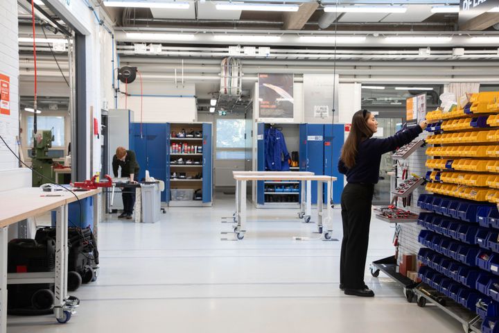 Ihminen järjestelee esineitä hyllyllä työtilassa, jossa on useita työkaluja ja kalusteita. Taka-alalla toinen henkilö työskentelee pöydän ääressä.