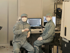 Kaksi tutkijaa puvuissaan OtaNano-laboratoriossa, pitelemässä pientä sirua monitorin edessä.