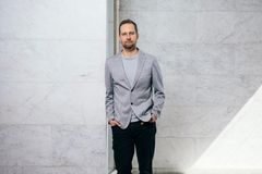 Henkilö seisoo vaaleaa kiviseinää vasten, pukeutuneena harmaaseen bleiseriin ja mustiin housuihin.