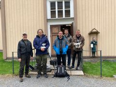 Viisi henkilöä seisoo ulkona vanhan puutalon edustalla kameravälineiden kanssa.