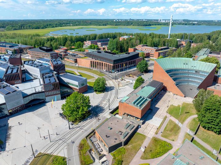 Ilmakuva kampusalueesta, jolla on useita tiilisiä ja modernisti muotoiltuja rakennuksia vehreässä ympäristössä. Taustalla näkyy järvi ja metsikköä.