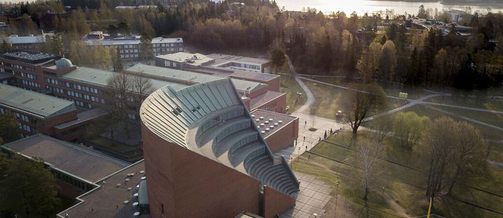 Ilmakuva Aalto-yliopiston kampusalueesta, jossa näkyy aaltomainen kattorakenne, kampusrakennuksia ja puustoista ympäristöä.