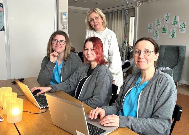 Kolme naista istuu pöydän ääressä tietokoneet edessään, heidän takanaan seisoo neljäs nainen.