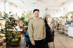 Mies seisomassa valoisassa avarassa kahvilatilassa, taustalla myyntitiski, pöytäryhmiä, viherkasveja.