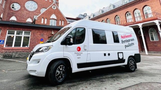 Punaisen tiilirakennuksen edessä seisoo valkoinen pakettiauto, jonka kyljessä on Diakonissalaitoksen logo ja teksti: "Rohkeasti ihmisarvon puolesta".