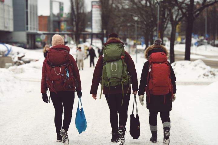 Kolme ihmistä reput selässä kävelevät kamerasta pois päin talvisessa kaupunkimaisemassa.