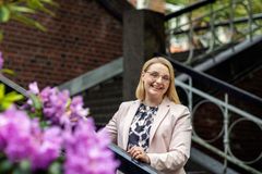 Vaaleaan bleiseriin pukeutunut nainen katsoo hymyillen kameraan. Taustalla punainen tiiliseinä ja vieressä kukkivia alppiruusuja.