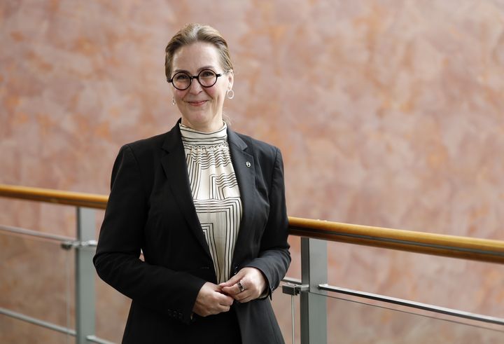 Sisätiloissa otettu valokuva henkilöstä, joka seisoo kaiteen läheisyydessä vaaleaa seinää vasten. Henkilöllä on yllään tumma bleiseri ja vaalea, kuviollinen paita sekä pyöreät silmälasit.